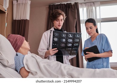 Caucasian Female Doctor In Uniform Diagnosis Explains X-ray Film With Asian Radiologist And Recovery Male Patient At Inpatient Room Bed In A Hospital Ward, Medical Clinic, Cancer Examination Consult.