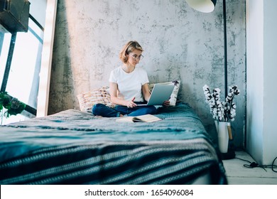 Caucasian female freelancer sitting at comfortable bed in cosy home interior and working remotely on modern laptop computer, skilled woman watching online webinar on website via netbook application - Powered by Shutterstock