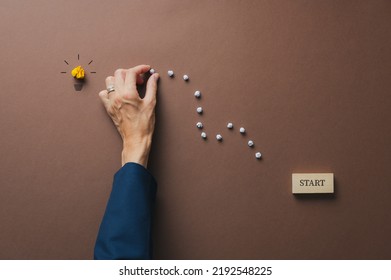 Caucasian Female Hand Making A Path From A Start Sign Towards A Light Bulb In A Conceptual Image Of Education, Ambition And Development. Over Brown Background.