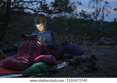 caucasian female hiker reading book/writing journal at night while wildcamping, strong light from headlamp