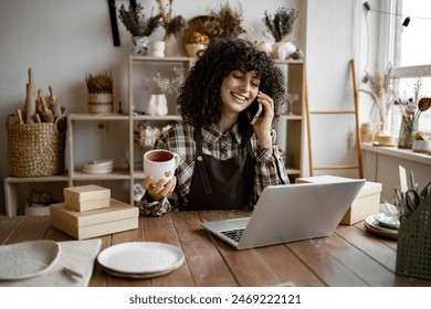 Caucasian female potter entrepreneur talking to buyer of handmade mug sitting at table and taking order using laptop. Successful business lady owner of shop made of clay talking to customer. - Powered by Shutterstock