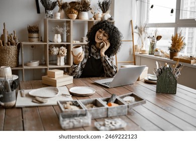 Caucasian female potter entrepreneur talking to buyer of handmade mug sitting at table and taking order using laptop. Successful business lady owner of shop made of clay talking to customer. - Powered by Shutterstock