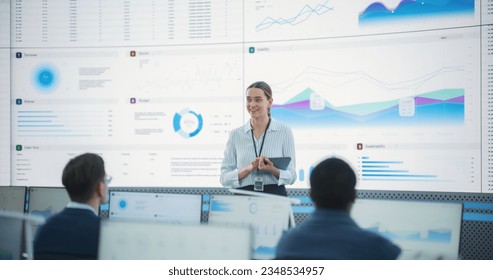 Caucasian Female Project Manager With Tablet Computer Is Giving Presentation To Diverse Employees In Front of Big Digital Screen In Monitoring Room. Team Of Data Scientists Listening To New Objectives