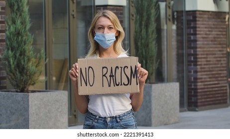 Caucasian Female Protester Activist Wearing A Medical Mask Holds A Cardboard Poster With The Slogan NO RACISM. A Protest Against Racism And Violence During Quarantine. Stop Racism Concept.