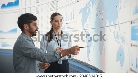Caucasian Female Regional Manager Talking To Indian Male Distribution Expert In Front Of Big Digital Screen With Infographics And Map. Successful Man And Woman Discussing New Markets For Expansion.