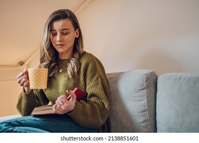 Caucasian female relaxing in a cozy living room while drinking tea and reading a book at home. Leisure weekend time with favorite novel alone at home.