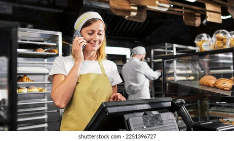 Caucasian female seller in bakeshop standing at paydesk and talking on mobile phone. Beautiful woman baker in apron and hat speaking on cellphone while selling bakery. Workday. Telephone call.