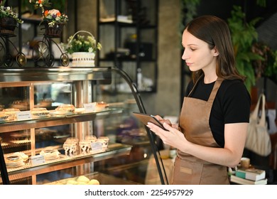 Caucasian female small business owner using digital tablet while standing near showcase with cakes and desserts