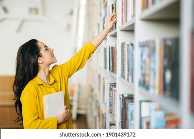 Caucasian female student stands in university library, looking for a book. Beautiful young woman in eyeglasses searching information for a project, learning for passing exams, education concept - Shutterstock ID 1901439172
