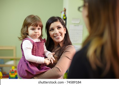 Caucasian Female Teacher And Mom With Baby Girl Talking In Kindergarten. Horizontal Shape, Focus On Background