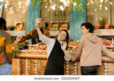 Caucasian female vendor holding mobile device, taking pictures with customers at farmers market for social media content. Happy small business owner promoting fresh organic fruits and vegetables. - Powered by Shutterstock