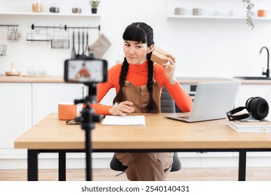 Caucasian female vlogger engages audience while unpacking product during live streaming session. Enthusiastic content creator communicates with followers using smartphone and laptop in kitchen setting - Powered by Shutterstock