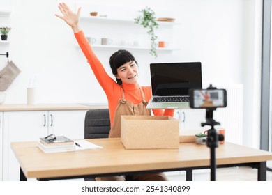 Caucasian female vlogger excitedly unboxes new digital laptop during online video recording at home office. Young woman shares unboxing experience with audience using smartphone camera setup. - Powered by Shutterstock