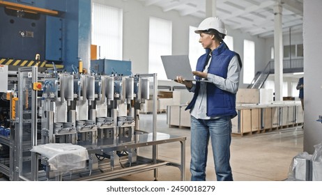 Caucasian female wearing vest and helmet for protection. Standing next to foil production line. Checking data while recording numbers on laptop. Man coming behind with digital device. - Powered by Shutterstock