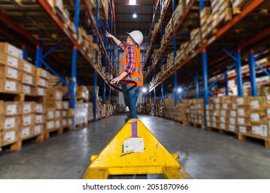 A Caucasian Female Worker Pulling A Pallet Truck In A Large Distribution Center. Woman Searching The Location Of Parcel On The Shelf For Loading The Boxes.