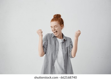 Caucasian freckled woman with red hair rejoicing her success and victory clenching her fists with joy. Lucky woman with hair bun being happy to achieve her aim and goals. Positive emotions, feelings