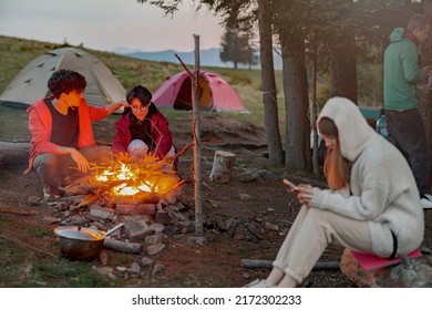 Caucasian Friends Hikers Warming Up At Fire In Camp Site In Mountains. Evening.