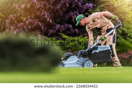 Caucasian Garden and Landscaping Worker Mowing  Backyard Lawn Using Electric Cordless Grass Mower. Garden Maintenance Theme.