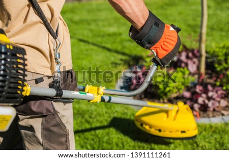 Caucasian Gardener with Gasoline Brush Cutter and String Trimmer on His Shoulder. Trimming Garden Grass. Landscape Maintenance.