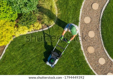 Caucasian Gardener Mowing Backyard Garden Grass Using Cordless Electric Grass Mower. Aerial View. Gardening and Landscaping Industry.