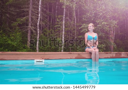 Caucasian girl in a blue swimsuit sits near the forest pool in the open. She looks away and smiles. The girl is on the right side of the frame. The concept of summer holidays, travel. Place for text.