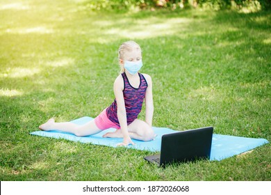 Caucasian Girl Child In Face Mask Doing Sport Workout Outdoor Online. Video Yoga Stretching On Internet. Kid Learning Training On Backyard With Laptop. New Normal. Social Distance At Coronavirus.