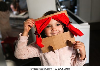 Caucasian Girl Child Plays In A Cardboard Theater With A Red Curtain, The Child Himself Makes Toys At Home, Children's Creativity