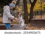 Caucasian girl holding a dog by the paws for a walk in the autumn park. Jack Russell Terrier stands on its hind legs on a bench.