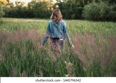 Caucasian girl with long brown hair wearing a denim jacket walking through a field in the evening sun with hands touching grass - Shutterstock ID 1445796542