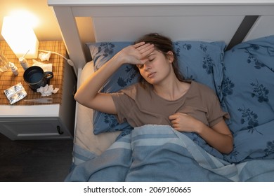 Caucasian Girl Lying In Bed, Touching Her Forehead, Suffering From Heat. Illness Cute Woman Tired Of A Long Illness. Female Got Sick With Flu, Cold, Angina Or Sore Throat. Top Down View. Healthcare.