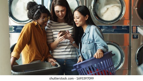 Caucasian Girl Showing Photos On Smartphone To Mixed-races Female Friends While Washing Machines Working And Cleaning Clothes. Multiethnic Women Watching Video On Phone In Laundry Service.