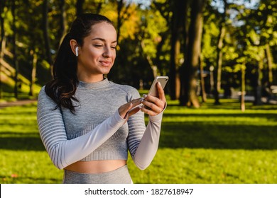 Caucasian girl in sport outfit is smiling and using her smart phone in a green park. - Powered by Shutterstock