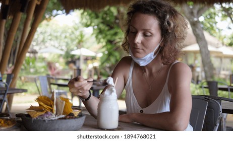 Caucasian Girl Takes Off Her Protective Face Mask To Try Whipped Cream Of Her Milkshake During Lunch At A Outdoor Restaurant. 