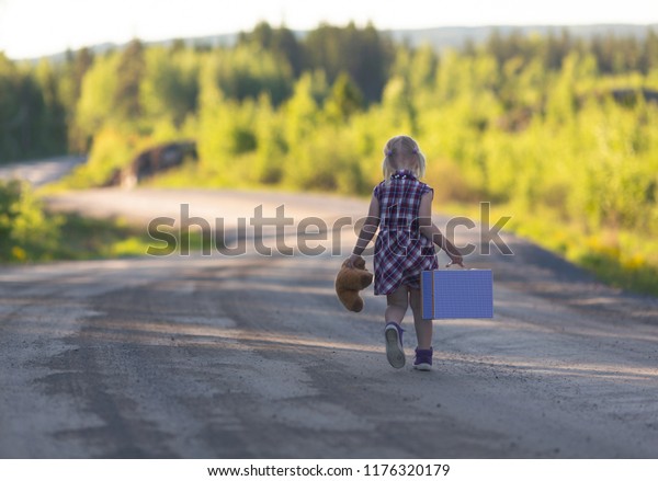 Caucasian girl walking away\
alone on an empty highway with bear and suitcase. Concept image of\
a runaway child with loneliness and sad feelings. Image has a\
vintage effect.
