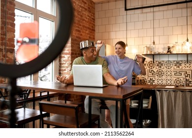 Caucasian Girl Want Hitting Pan On Head Of Black Guy Wearing Saucepan At Home Kitchen. Smiling Multiracial Blogger Couple Making Video In Prank Or Challenge Style On Mobile Phone For Social Networks