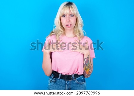 Caucasian girl wearing pink T-shirt isolated over blue background keeps hands on chest feeling shocked and scared, mouth widely opened, stares at camera saying: Who, me?