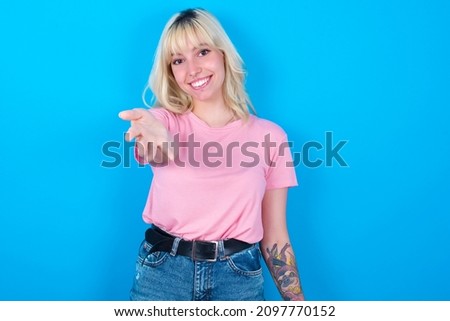 Caucasian girl wearing pink T-shirt isolated over blue background  smiling friendly offering something with open hand or handshake as greeting and welcoming. Successful business.