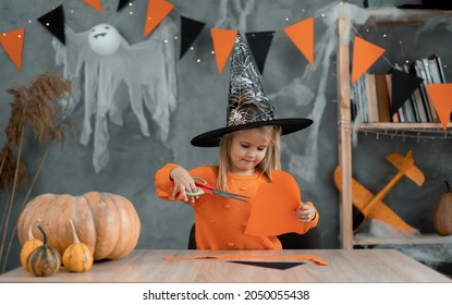 Caucasian Girl In A Witch Costume Sits At The Table At Home And Prepares Handicrafts For The Halloween Holiday. Diy And Decor In The Room Made Of Paper.