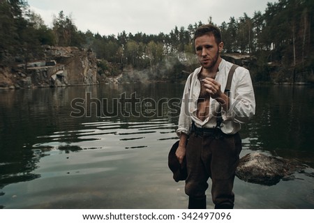 caucasian gold digger in retro clothes smokes cigarette while standing in lake with rocky bank after hard work. He wears shirt, leather pants, galluses and boots.  Sky is cloudy and grey.  Stock photo © 