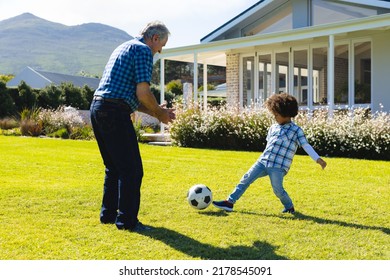 Caucasian Grandfather Playing Soccer With Biracial Grandson On Grassy Land In Yard On Sunny Day. Family, Unaltered, Togetherness, Childhood, Lifestyle, Retirement, Enjoying And Sport Concept.