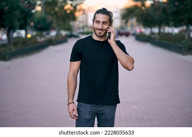 caucasian guy black t-shirt blue pants with beard smiling standing on the street talking on his smartphone on the city boulevard - Shutterstock ID 2228035633