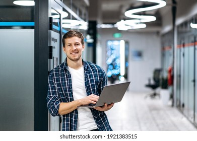 Caucasian guy freelancer with laptop. A successful guy dressed in stylish casual wear stands in a creative office, holds a laptop, looks and smiles friendly at the camera