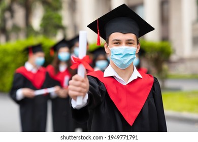 Caucasian Guy Student In Graduation Costume And Protective Face Mask Showing His Diploma At Camera, Posing Over International Group Of Students At University Campus, Selective Focus, Copy Space