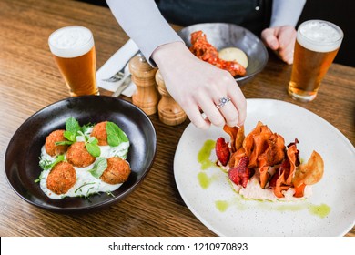 Caucasian hand grabbing vegetable chips in hummus on a white plate next to a black bowl of croquettes in tzatziki next to a pint of beer on a wooden table.