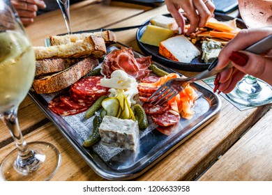 Caucasian Hand Holding A Fork Next To A Platter Of Deli Meat, Bread And Pickled Vegetables On A Silver Platter On A Wooden Table.