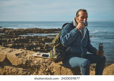 Caucasian handsome hiker traveler man sitting on the cliff and enjoying drinking hot drink. Navigational equipment lying down on the rock. People. Travel. Nature - Powered by Shutterstock