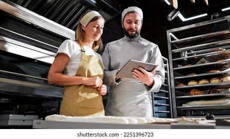 Caucasian handsome man baker in uniform showing something on tablet device to woman colleague. Bakers talking and discussing work with computer in hands. Bakery concept. Workday at bakeshop.