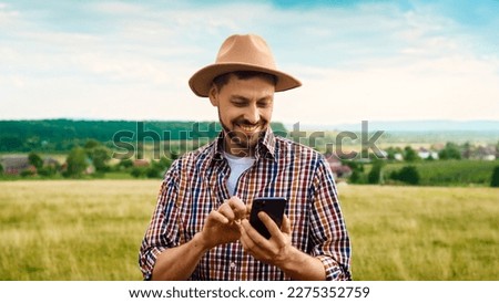 Caucasian handsome man in hat standing in grassland and using mobile phone. Outside. Good-looking young male farmer tapping and texting on smartphone. Gadget for farming. Chatting while working.
