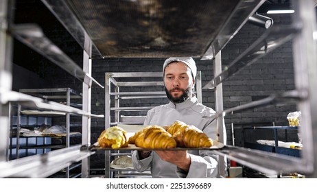 Caucasian handsome man in hat and uniform working in bakehouse and putting croissants on shelf. Male baker making fresh pastries in kitchen of bakeshop in morning. Confectionery producing concept. - Shutterstock ID 2251396689