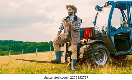 Caucasian handsome young man farmer in hat standing at tractor, using smartphone and resting in field. Countryside worker concept. Happy male having rest and texting on phone while chatting. Outdoor.